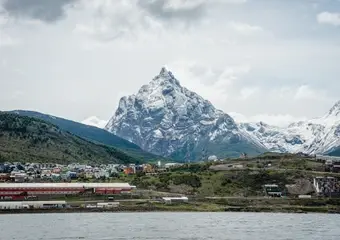 Spectaculair uitzicht op een imposante besneeuwde bergpiek boven een kleurrijke havenstad aan het water, met groene hellingen en industriële infrastructuur in de voorgrond.