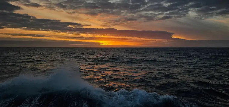 Een dramatische zonsondergang boven de oceaan met gouden en oranje tinten aan de hemel, terwijl donkere golven met wit schuim tegen het strand breken.