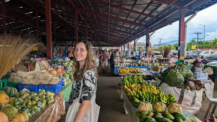Een vrouw poseren op een kleurrijke tropische markt onder een dak, omgeven door verse groenten, fruit en kruiden, met andere bezoekers en handelaren op de achtergrond.