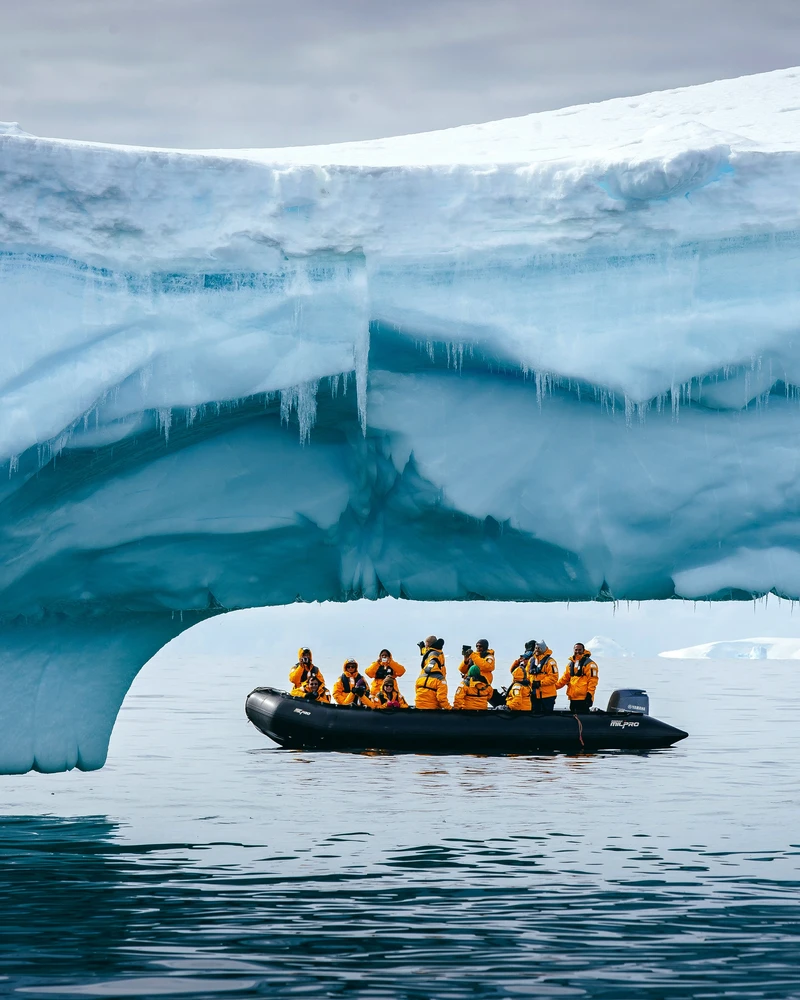 Een groep toeristen in gele reddingsvesten vaart met een zwarte rubberboot door een spectaculaire ijsgroeve onder een gigantische blauwe gletsjerwand met hangende ijspegels.