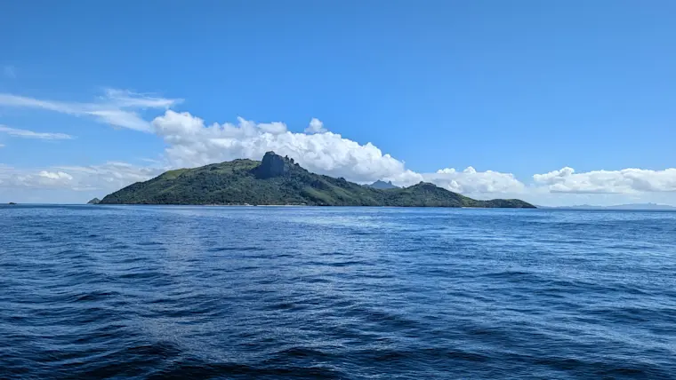 Een idyllisch tropisch eiland met weelderige groene begroeiing en karakteristieke bergtoppen, omgeven door kristalhelder blauw oceaanwater onder een stralende blauwe hemel met witte wolken.