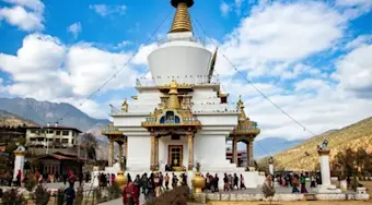 Een prachtige witte boeddhistische stupa met goudenDetailswerk omgeven door bergen, waar toeristen en lokale bezoekers bijeenkomen in een serene bergomgeving.