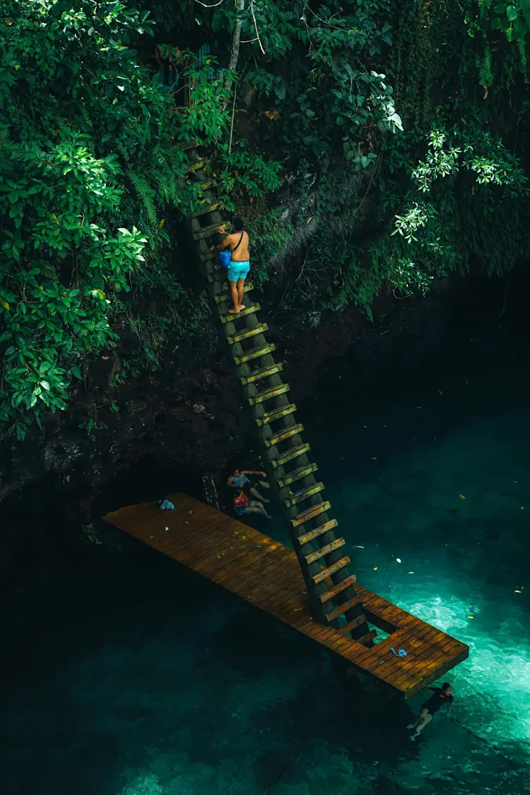 Een avonturier klimt via een houten trap langs een met groen begroeide rots naar het kristalhelder turquoise water in wat lijkt te be een verborgen tropische grot of cenote.