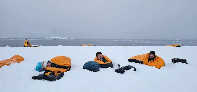 Groep van mensen in gele reddingsjassen liggend op het ijs tijdens een poolexpeditie, met ijsschotsen en water op de achtergrond.