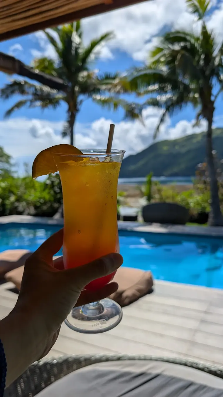 Een tropische cocktail wordt vastgehouden aan de rand van een zwembad met uitzicht op palmen, bergen en een idyllisch eilandlandschap onder een blauwe hemel.