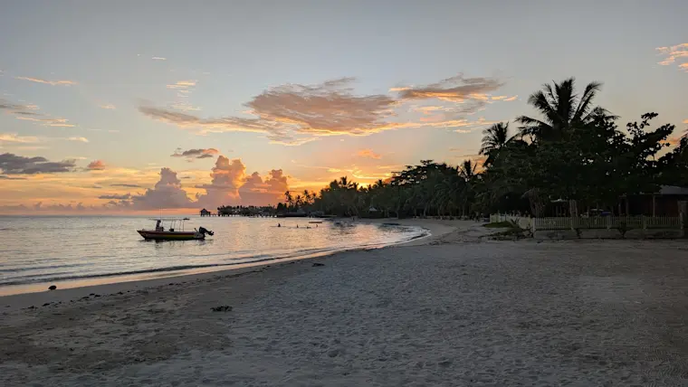 Een serene zonsondergang aan een tropisch strand met palmbomen, een traditionele vissersboot in het ondiepe water en een rustige baai omgeven door groene vegetatie.