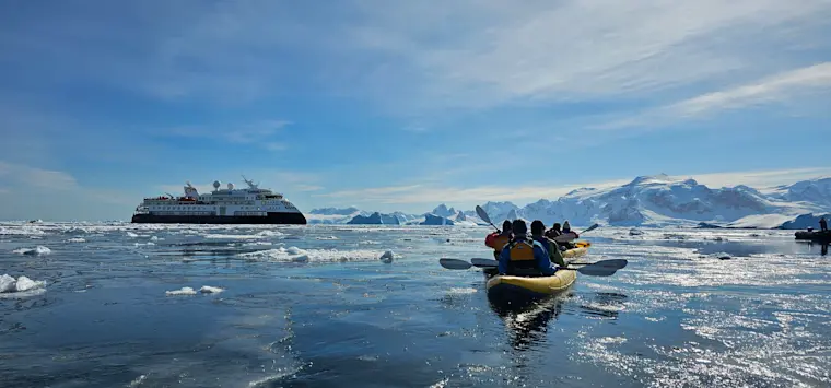 Een groep toeristen in een rubberboot verkent een ijzig polaire landschap met een cruiseschip op de achtergrond, omgeven door drijvend ijs en sneeuwbedekte bergen.