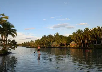 Een idyllisch tropical waterkanaal met palmbomen, traditionele houten huisjes aan weerszijden en een stand-up paddler op het kalme water onder een zonsonderganghemel.