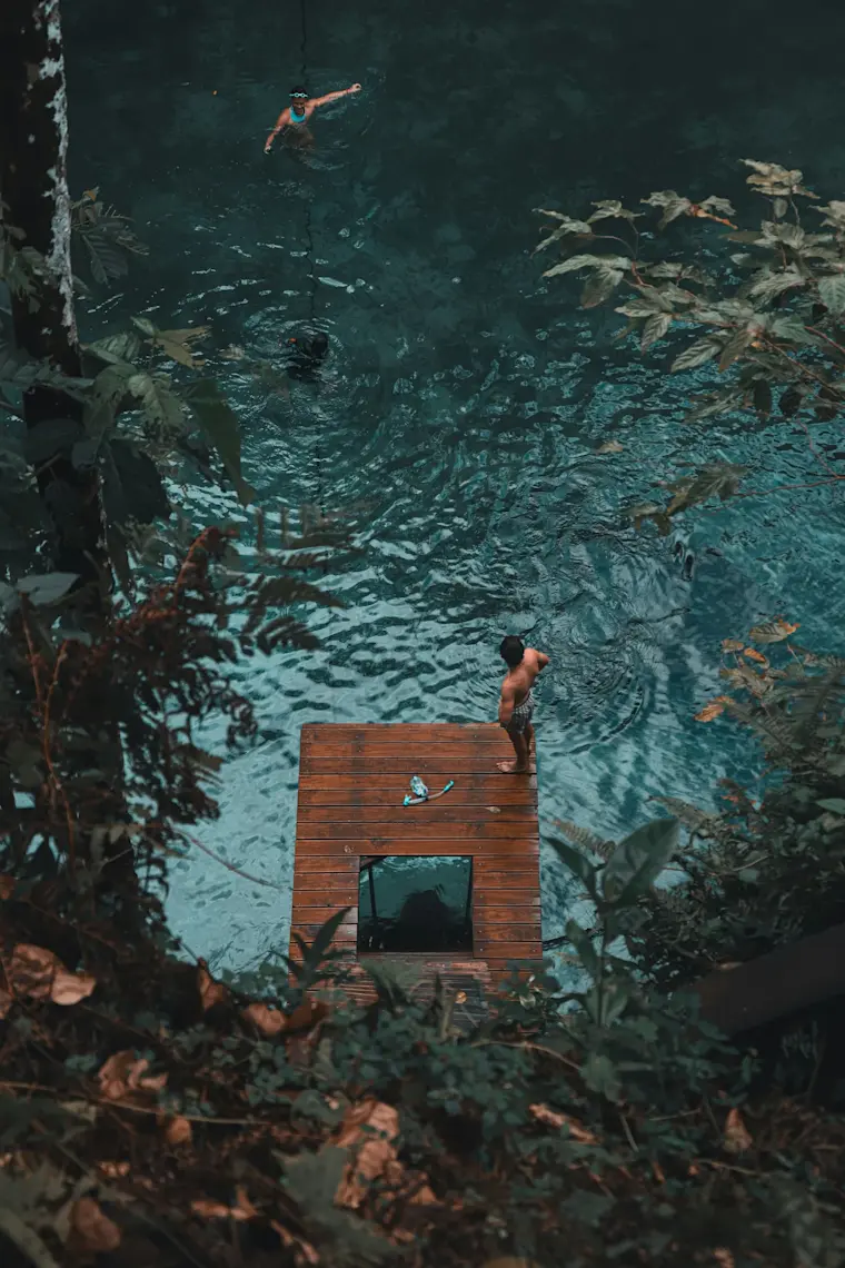 Luchtfoto van een houten platform aan een turquoise water omgeven door tropische vegetatie, waar zwemmers en duikers genieten van een exotische badplek.