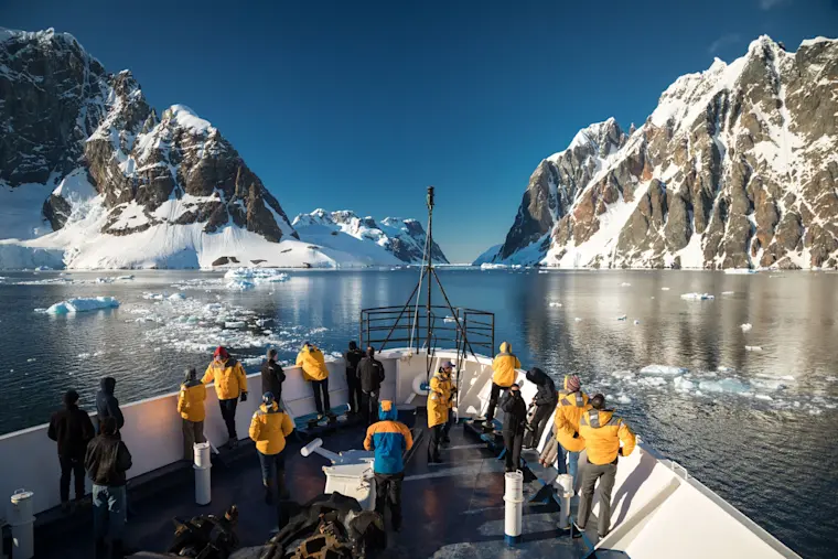 Toeristen in gele jassen aan boord van een expeditieboot in een besneeuwde Antarctic baai met imposante ijsbergen en rotswanden.