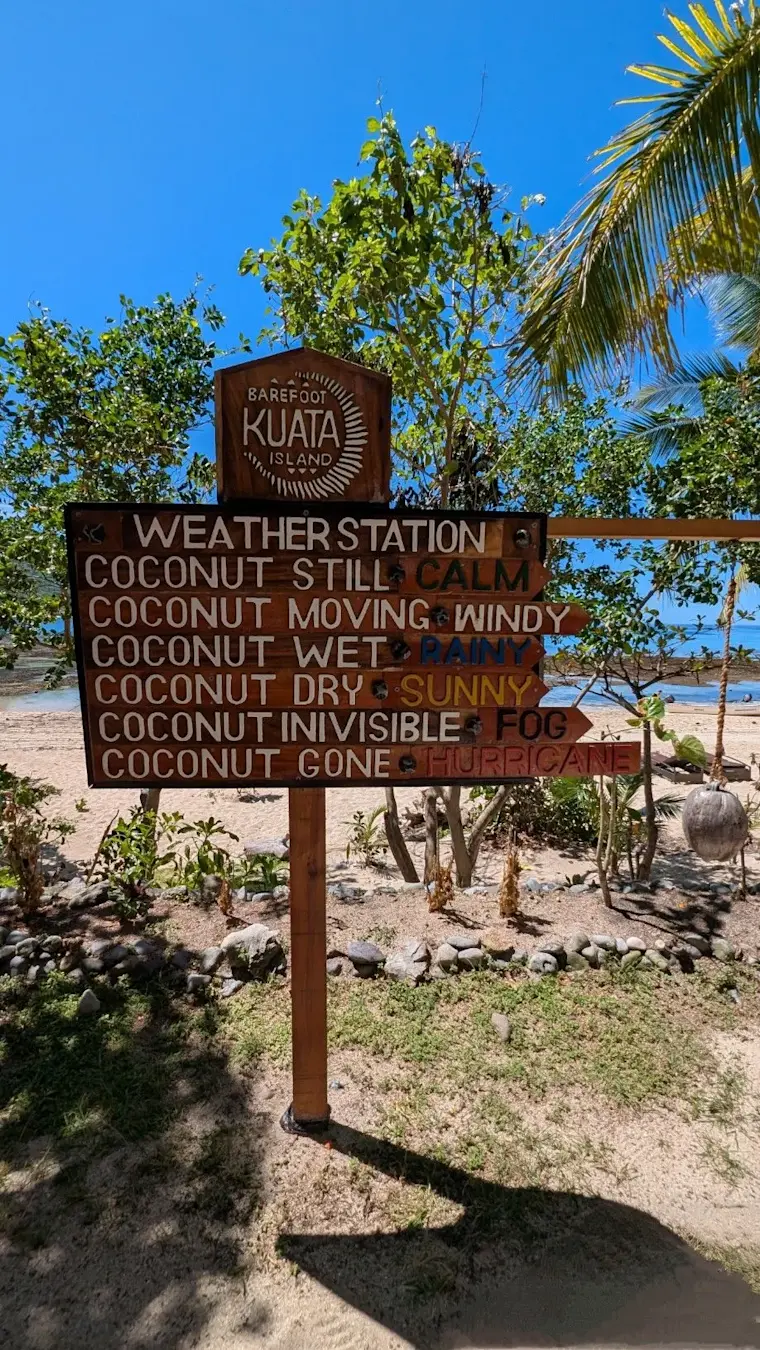 Een humoristische houten weerkaart op het eiland Kuata met grappige weerbeschrijvingen geformuleerd als kokosnoot-toestanden, geplaatst op een tropisch strand met palmen en blauwe hemel.