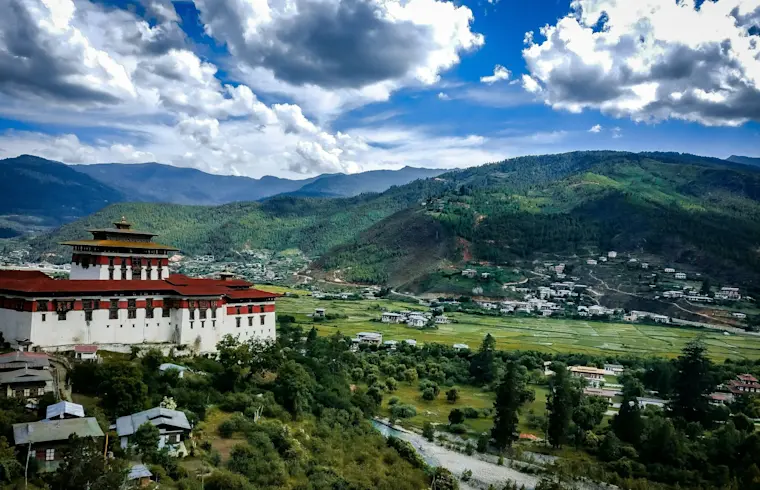 Het majestueuze Rinpung Dzong fort domineert het groene valle van Paro in Bhutan, omgeven door traditionele huizen, beboste bergen en een kristalhelder rivierje onder een wolkenloze blauwe hemel.