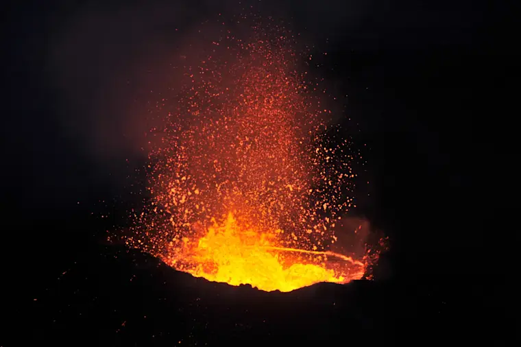 Een spectaculaire nachtfoto van een actieve vulkaan met gloeiende lava die hoog de lucht in spuit, omgeven door vonken en oranje-rode vuurstralen tegen een donkere nachtelijke hemel.