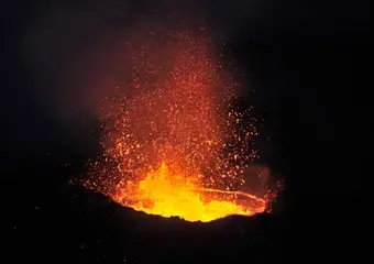 Een spectaculaire nachtfoto van een actieve vulkaankrater met gloeiende lava en vuurwerk van gesmolten gesteente dat hoog de lucht in wordt geslingerd tegen een donkere sterrenhemel.