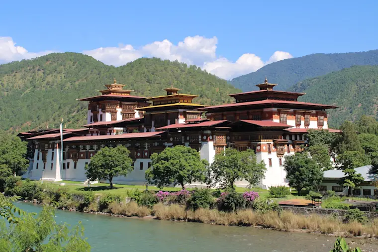 Het iconische Tashichho Dzong, een prachtig traditioneel Bhutaans klooster-fort met karakteristieke bruine en witte architectuur, gesitueerd aan de oever van een turquoise rivier omgeven door dichte groene bergen.