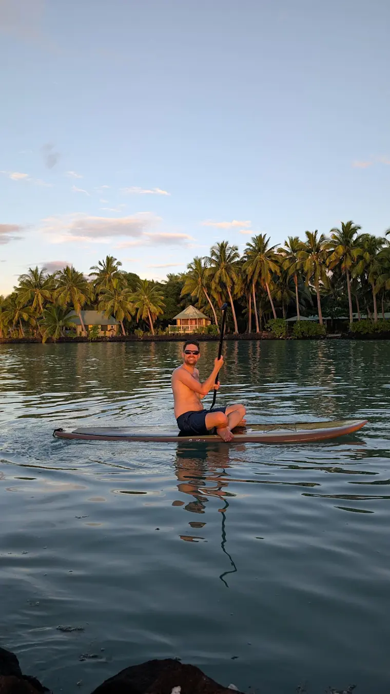 Een man zit op een surfboard in het water voor de kust van Fassad Village, met op de achtergrond palmbomen en traditionele huizen aan de rivier tijdens een prachtige zonsondergang.