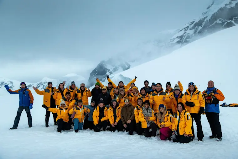 Een grote groep toeristen in gele en blauwe expeditiejassen poseert voor een groepsfoto op een besneeuwde gletsjer met imposante besneeuwde bergtoppen op de achtergrond.