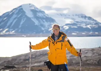 Een glimlachende wandelaar in gele bergjas met trekkingstokken poseert voor een adembenemend landschap met sneeuwbedekte vulkanen en een bevroren meer in wat lijkt te zijn IJsland of een soortgelijke arctische bestemming.