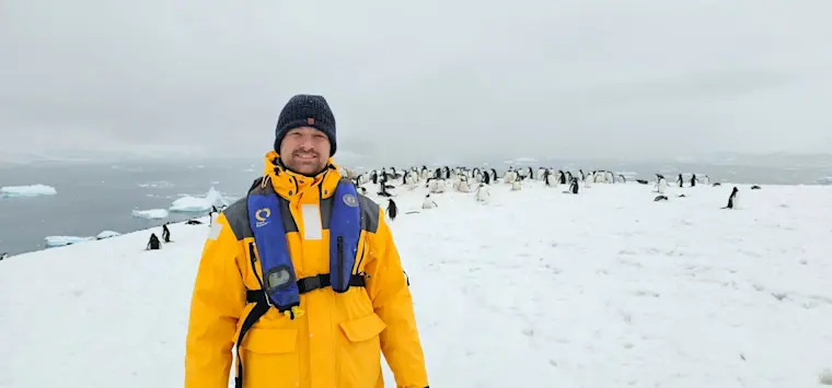 Een avonturier in gele expeditiejas poseert op een besneeuwde ijsberg omgeven door pinguïns in Antarctica.