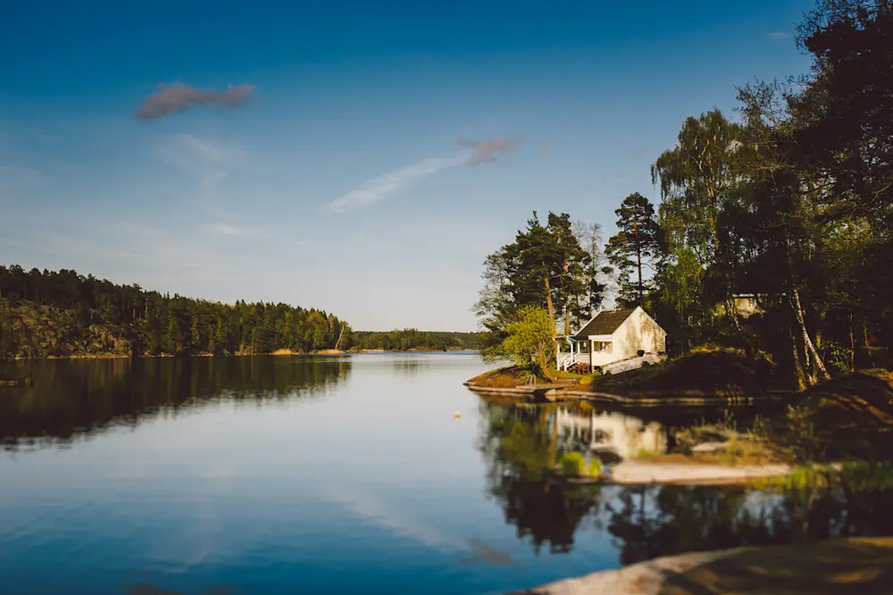 Fritidshus vid sjö med spegelblankt vatten, omgiven av skog och natur i lugn boendemiljö.
