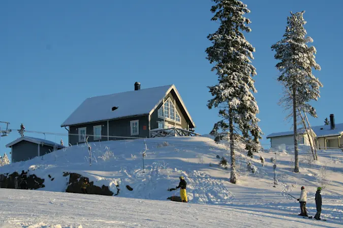 Fritidshus i snötäckt fjällmiljö med skidåkare i backen och fjällbjörkar runt huset.