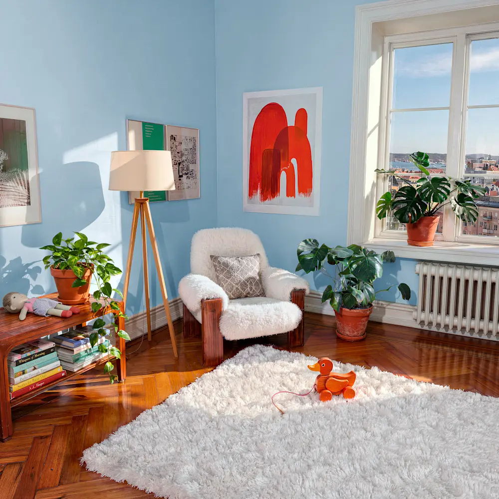 Ljust vardagsrum med blå väggar, fåtölj, växter, stor matta och fönster med utsikt över staden.