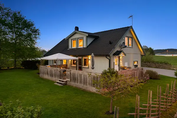 Villa med träfasad, stor altan och trädgård i kvällsljus, omgiven av grönska i lugnt bostadsområde.