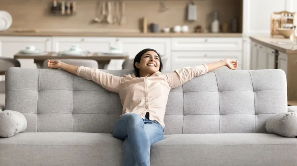 Person sitter utsträckt i grå soffa i vardagsrum med öppet kök i bakgrunden och ljus, modern inredning.