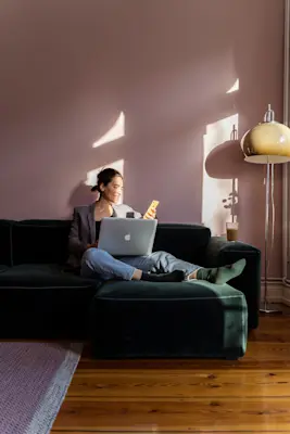 Person sitter i solbelyst vardagsrum på mörk soffa med laptop och mobil bredvid en golvlampa.