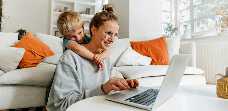 En kvinna sitter vid laptop i ljust vardagsrum medan ett barn klättrar upp bakom soffan.