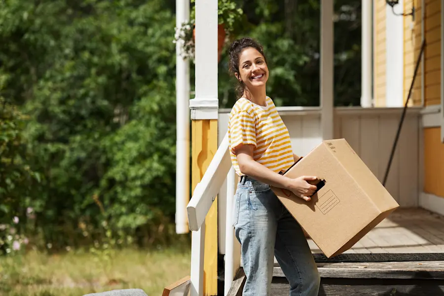 Person bär en flyttkartong på en veranda vid ett gult hus omgivet av grönska.
