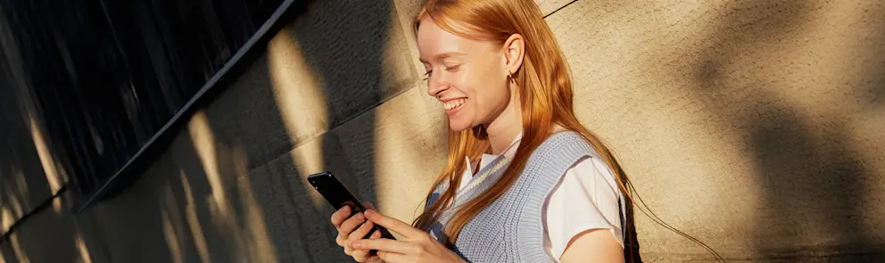 Person använder mobiltelefon utomhus i kvällssol mot byggnadsfasad i modern stadsmiljö.