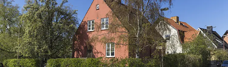 Rött hus med vita fönster omgivet av träd och häckar i ett villaområde under klar blå himmel.