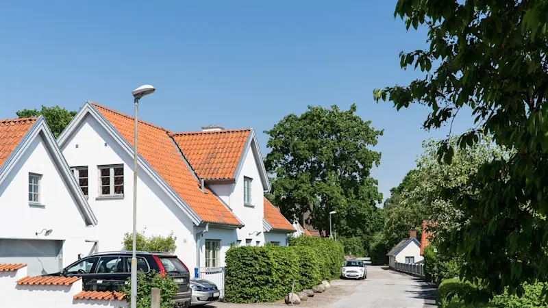 Villor med vita fasader och tegeltak längs lugn gata med grönska och parkerade bilar i bostadsområde.
