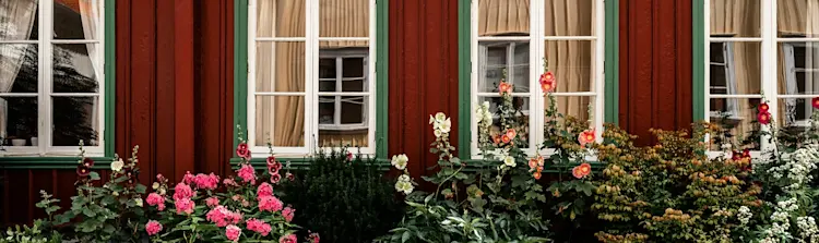 Röd träfasad med spröjsade fönster och blommande trädgård utanför ett traditionellt svenskt hus.