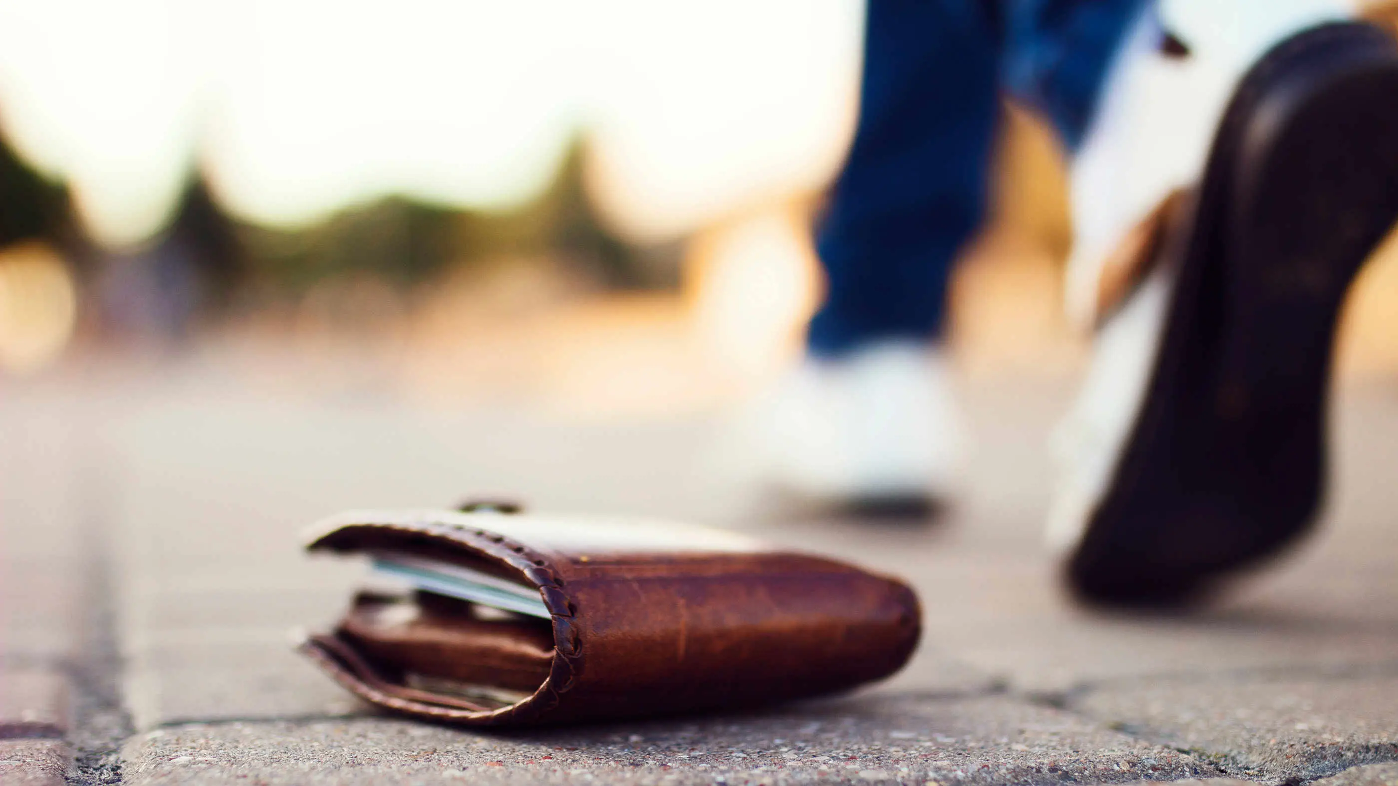 Ein verlorenes gefülltes Portemonnaie liegt auf dem Gehsteig, daneben sieht man die Füße einer vorbei gehenden Person.
