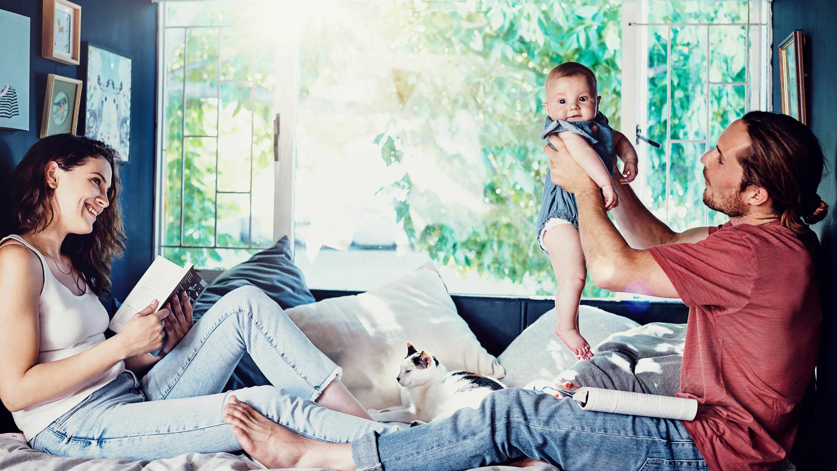 Ein Pärchen aus Mann und Frau sitzt gemeinsam mit ihrem Baby auf der Couch. Der Mann hält das Kleinkind in die Höhe.