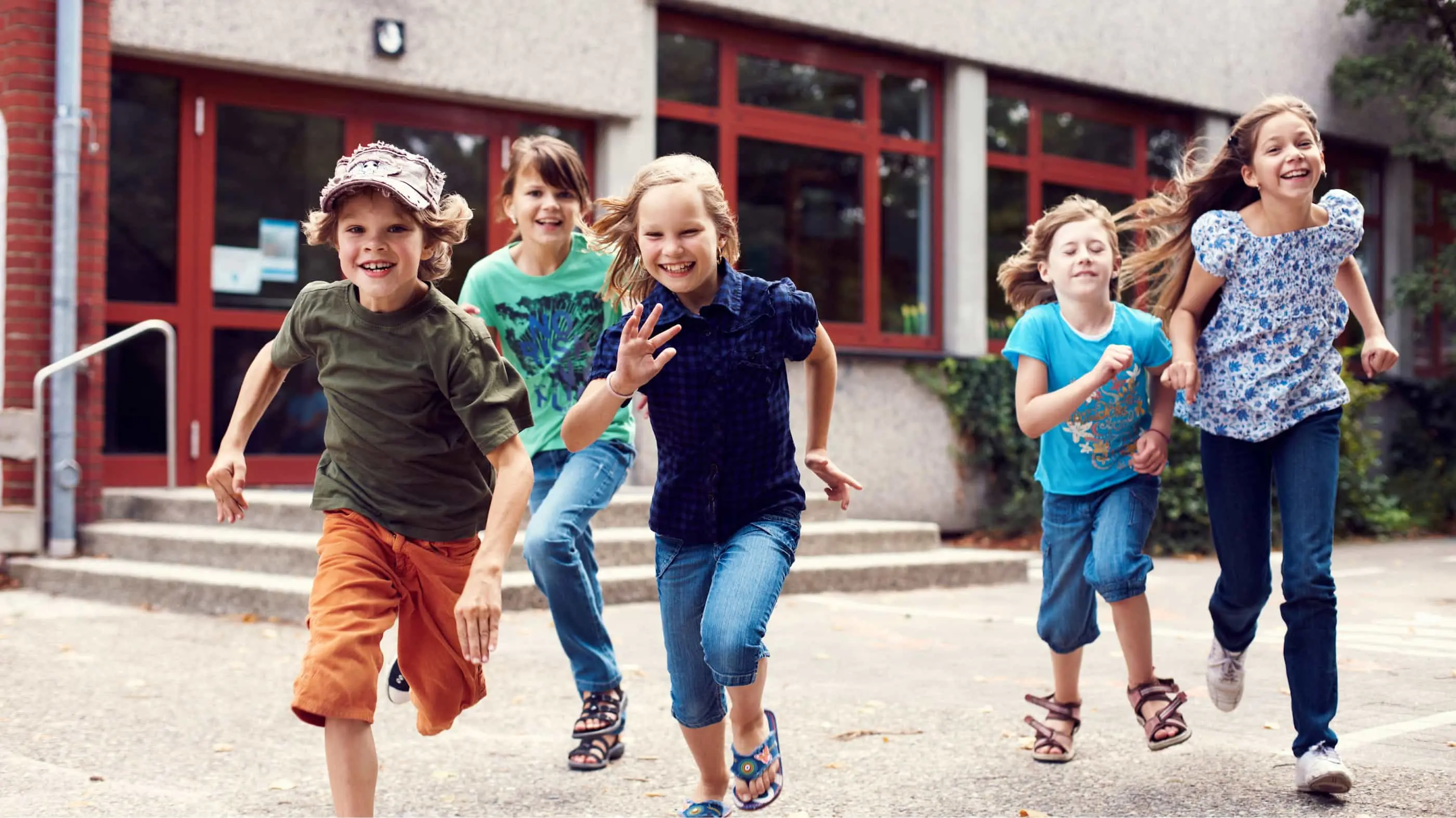 5 Kinder rennen fröhlich vom Eingang einer Schule oder Kita auf den Hof.