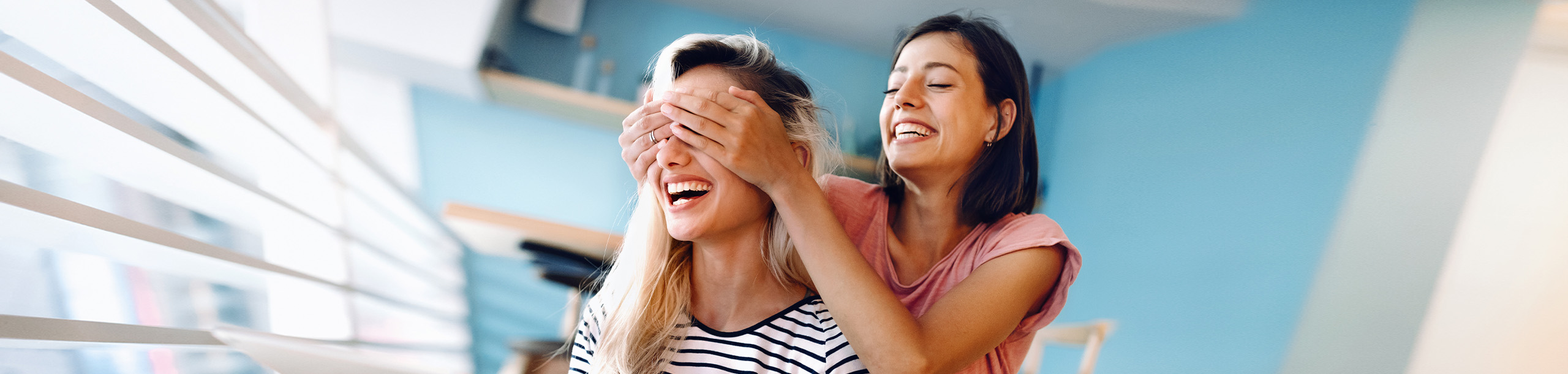 Eine Frau hält einer anderen Frau die Augen zu während sie lachend vor einem Tablet sitzen