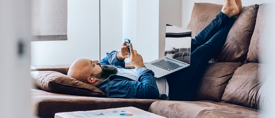 Ein Mann liegt mit Smartphone und Laptop auf der Couch