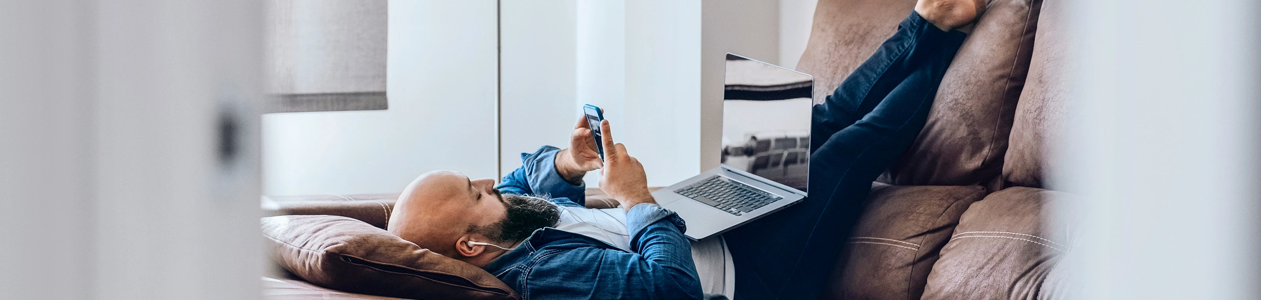 Ein Mann liegt mit Smartphone und Laptop auf der Couch