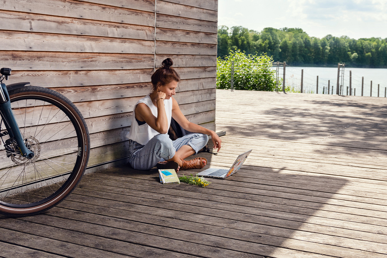 Frau sitzt am See mit Laptop und E-bike.