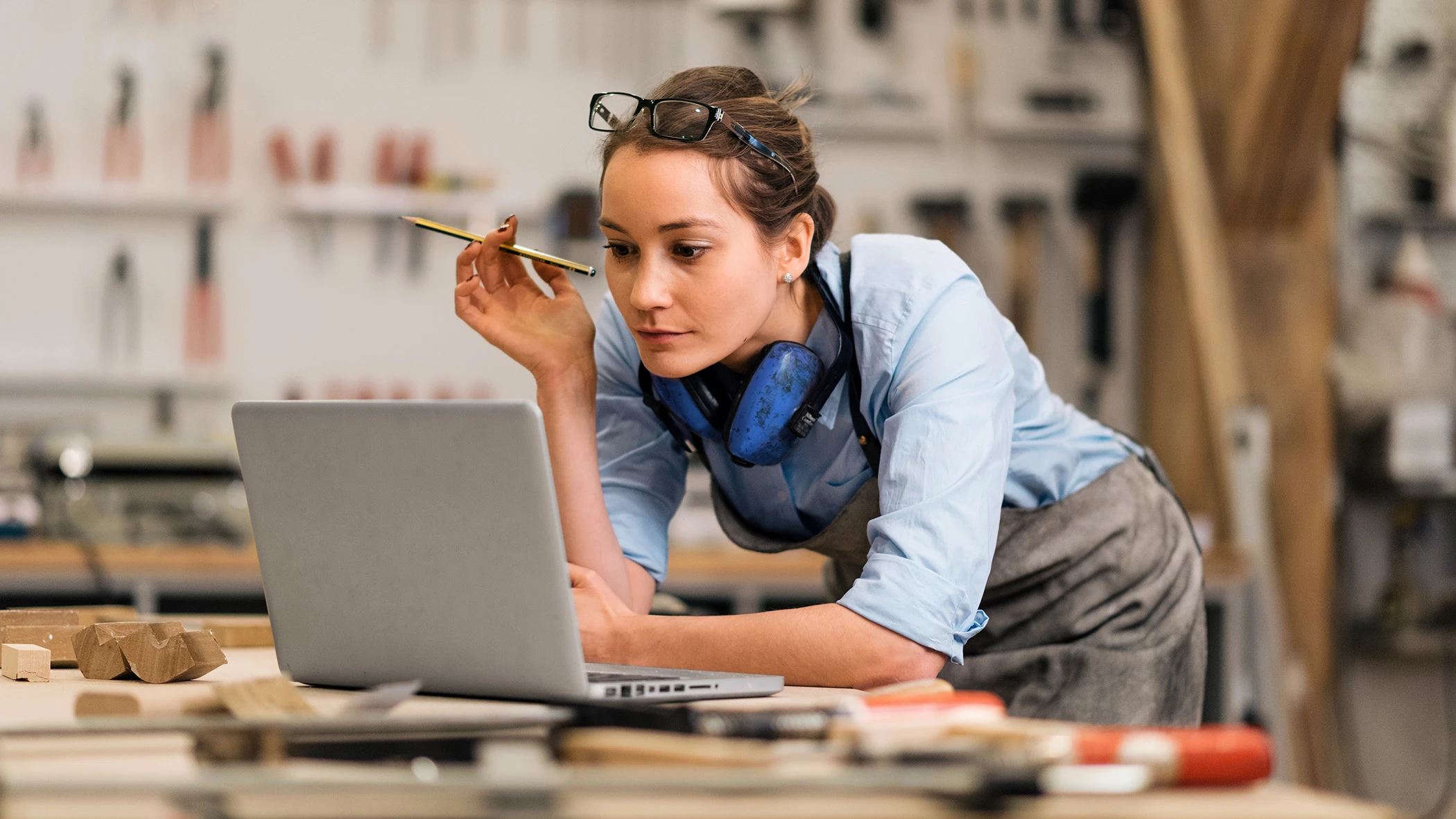 Eine Frau lehnt an einer Werkbank und schaut auf einen Laptop.