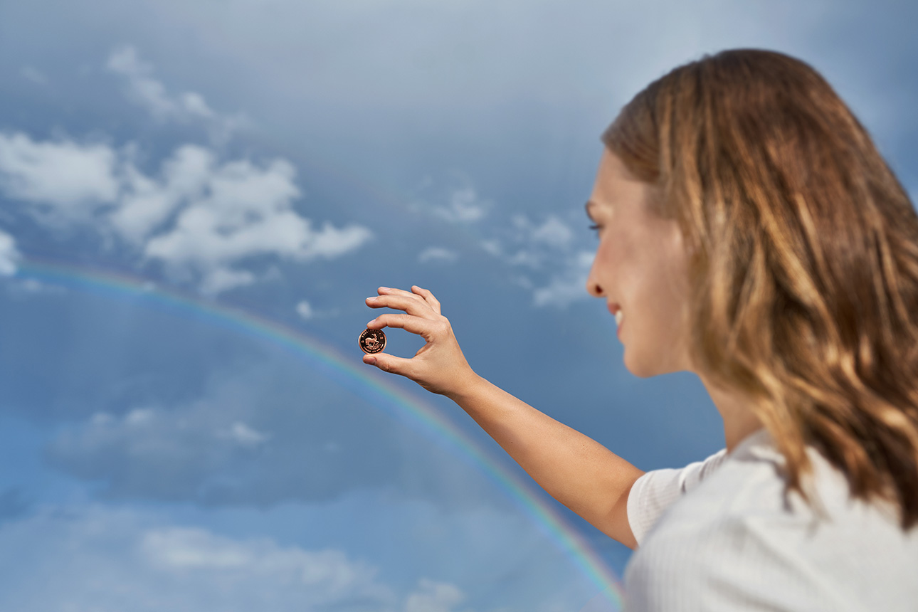 Frau betrachtet Goldmünze vor einem Regenbogen