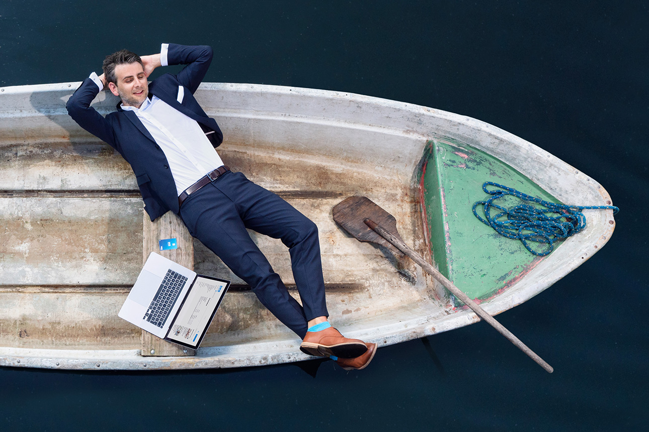 Ein Mann im Anzug liegt entspannt auf einem Boot, neben ihm ein aufgeklappter Laptop und eine Visa Kreditkarte.