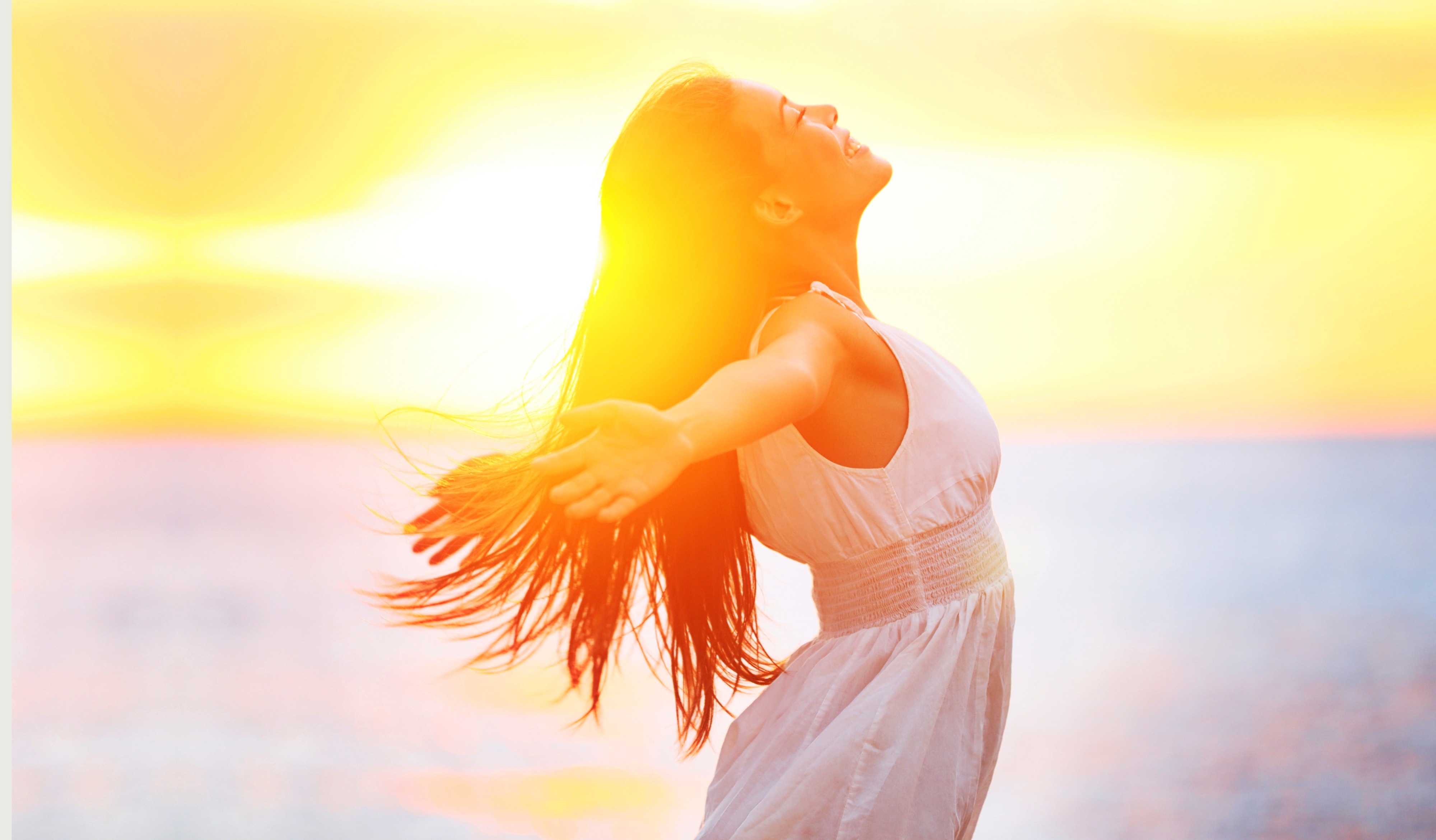 Nainen hymyilee kädet levitettynä, taustalla auringonlasku meren äärellä