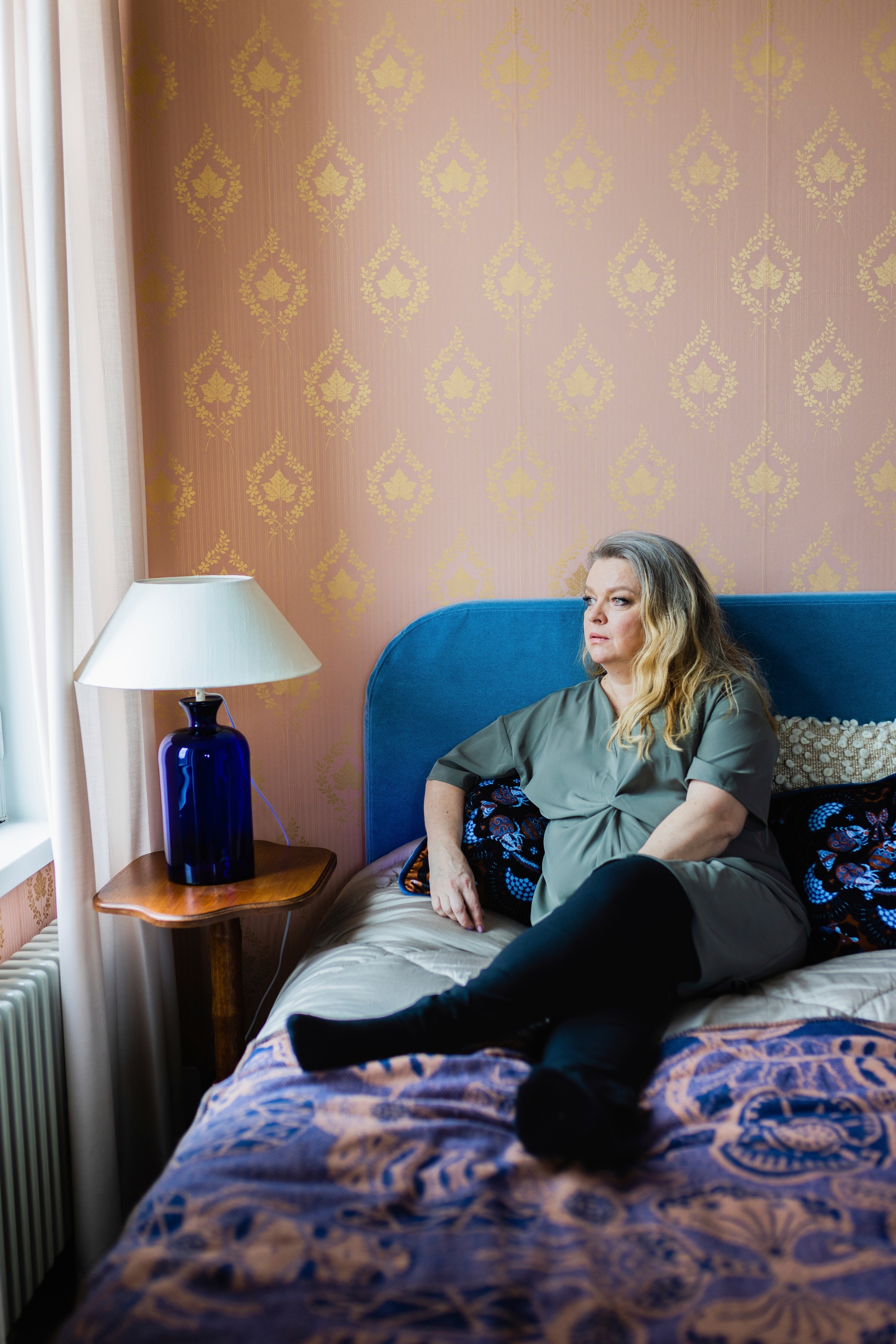 Elina Knihtilä makaa puoli-istuvassa asennossa sängyllä, katsoo ulos viereisestä ikkunasta