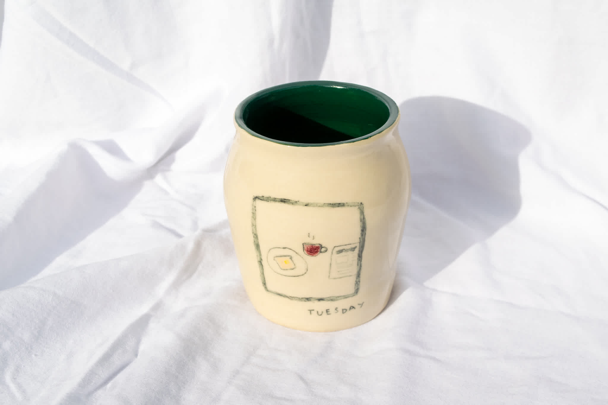 Cream ceramic exterior/green interior vase with drawing of toast, coffee, and newspaper. Placed on a white sheet