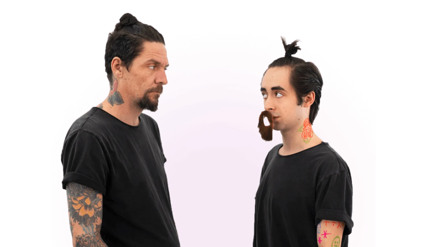 A man with a goatee, man bun, and tattoos on a black shirt stares at another person who is impersonating him by having the same tattoos, hairstyle, and t-shirt.