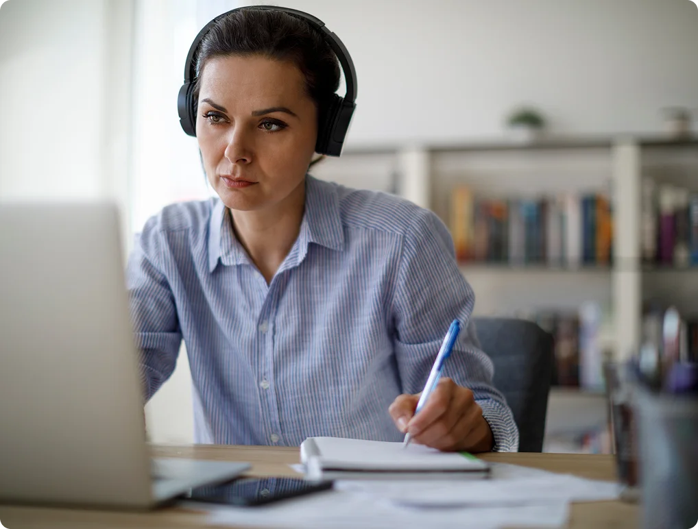 Professional with headset taking notes during virtual meeting at home office.